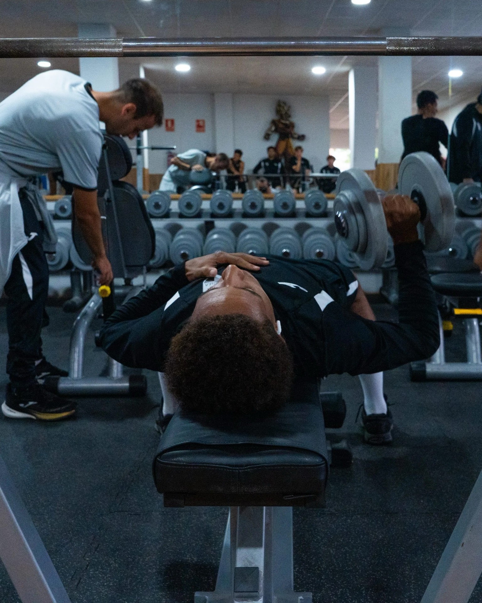 Un jugador hace press con mancuernas tumbado en un banco del gimnasio colaborador, con un preparador a su lado