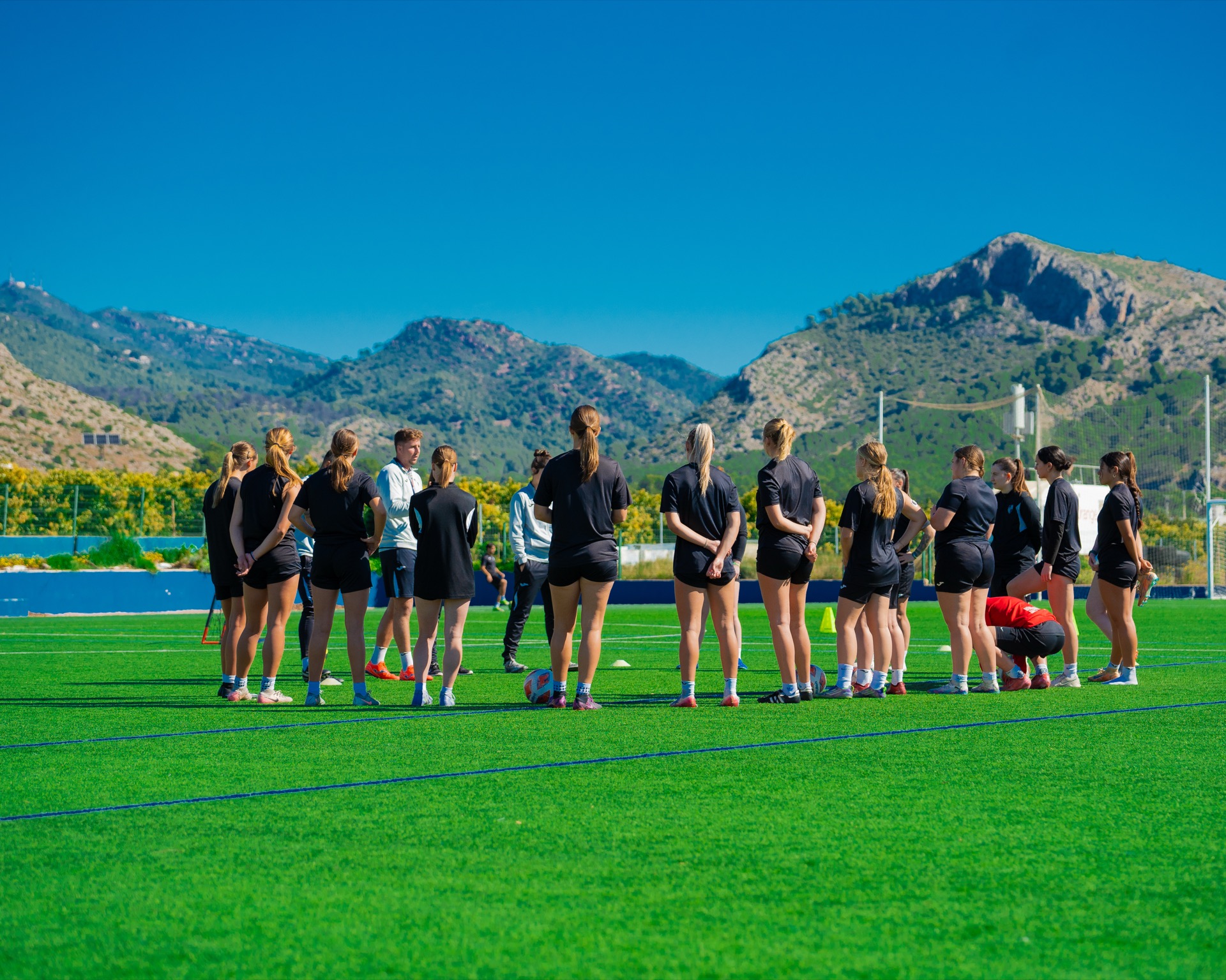 Las jugadoras de la academia femenina, en corro con el cuerpo técnico durante un entrenamiento, con las montañas al fondo