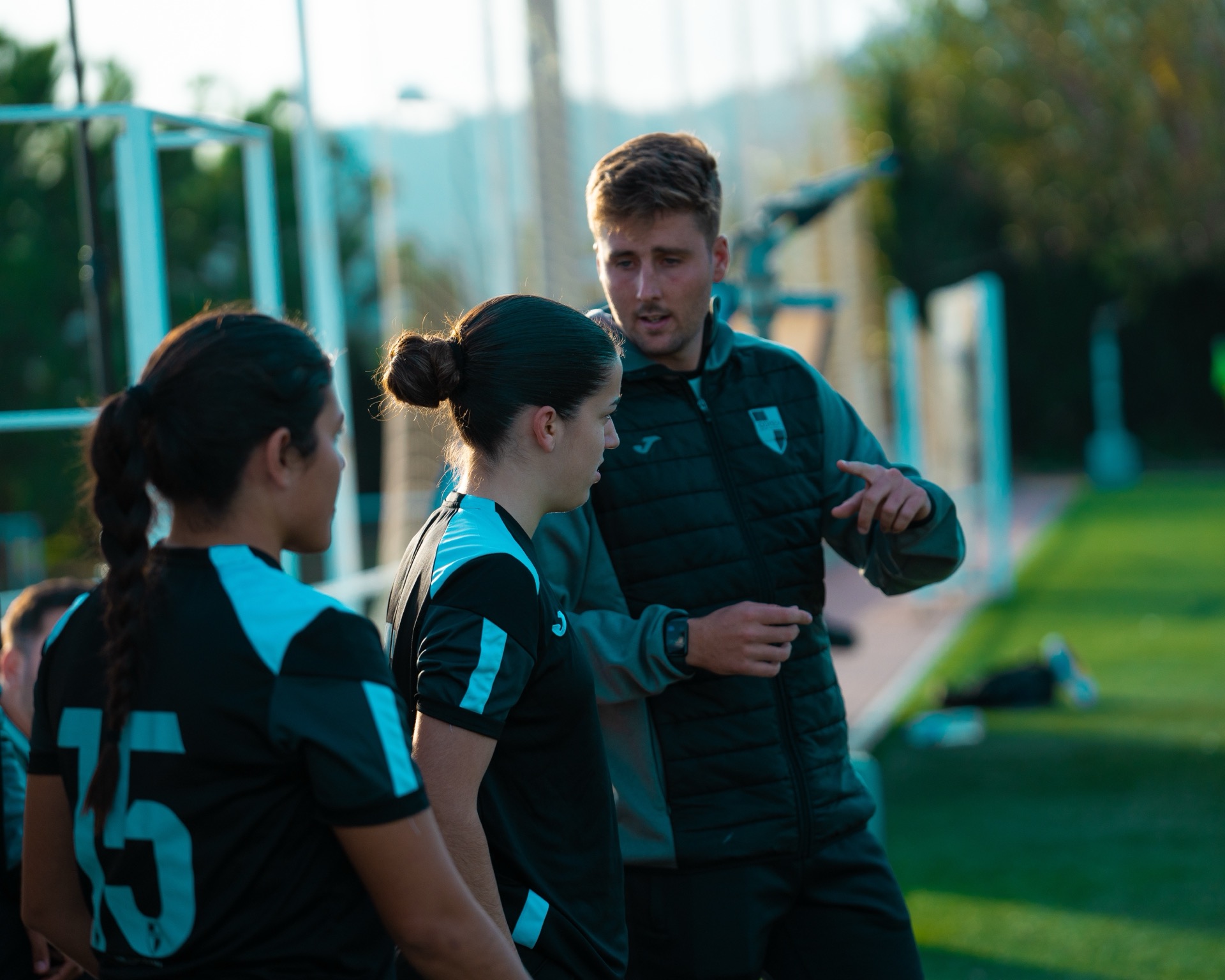 Un entrenador da indicaciones a dos jugadoras de la academia femenina, junto al campo