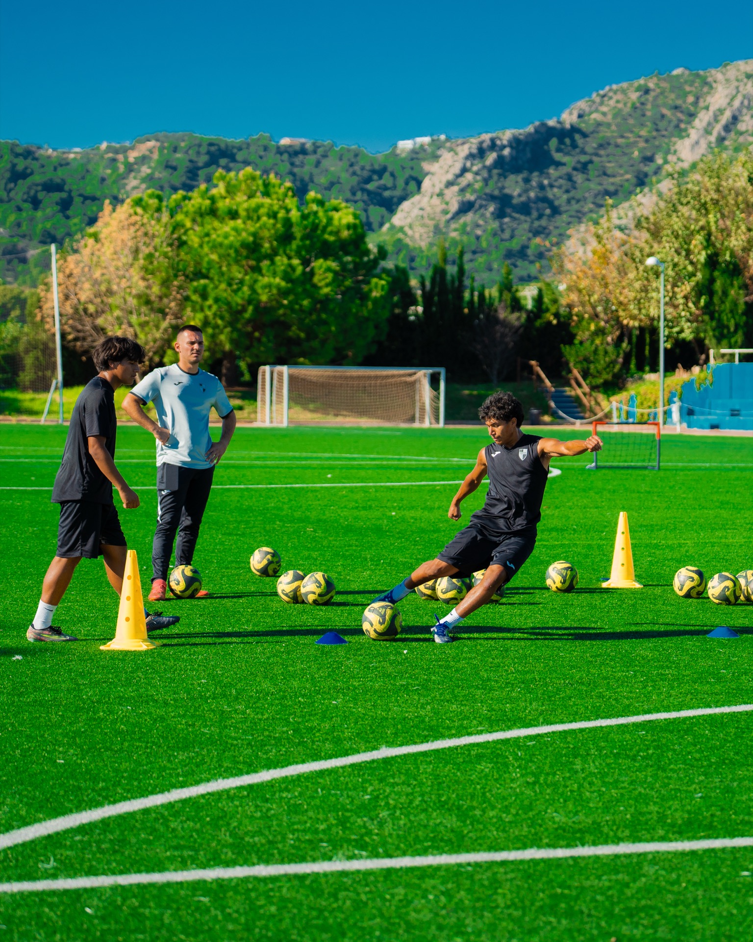 A player striking a ball past cones in a training drill, with a coach looking on